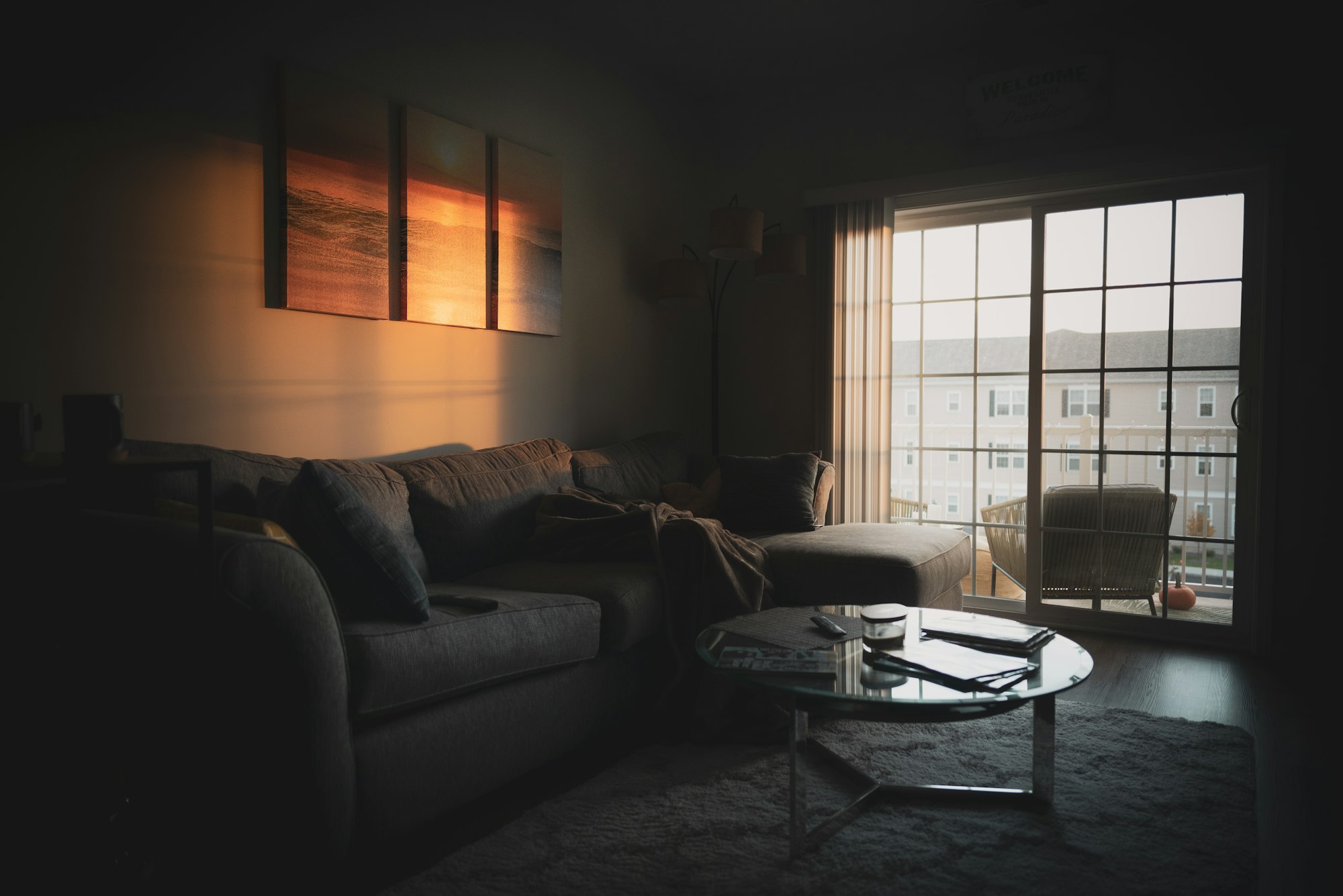 A dimly lit living room at dusk, with a grey sectional sofa, a round glass coffee table, and warm light falling across the wall.