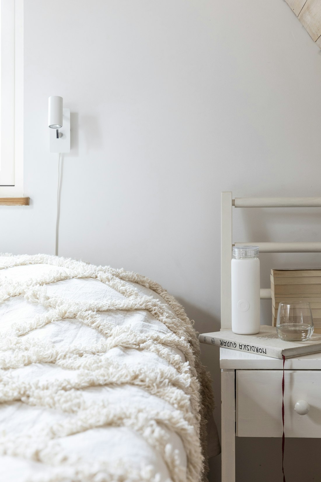 A textured cream bedspread beside a painted bedside table holding books and a glass.