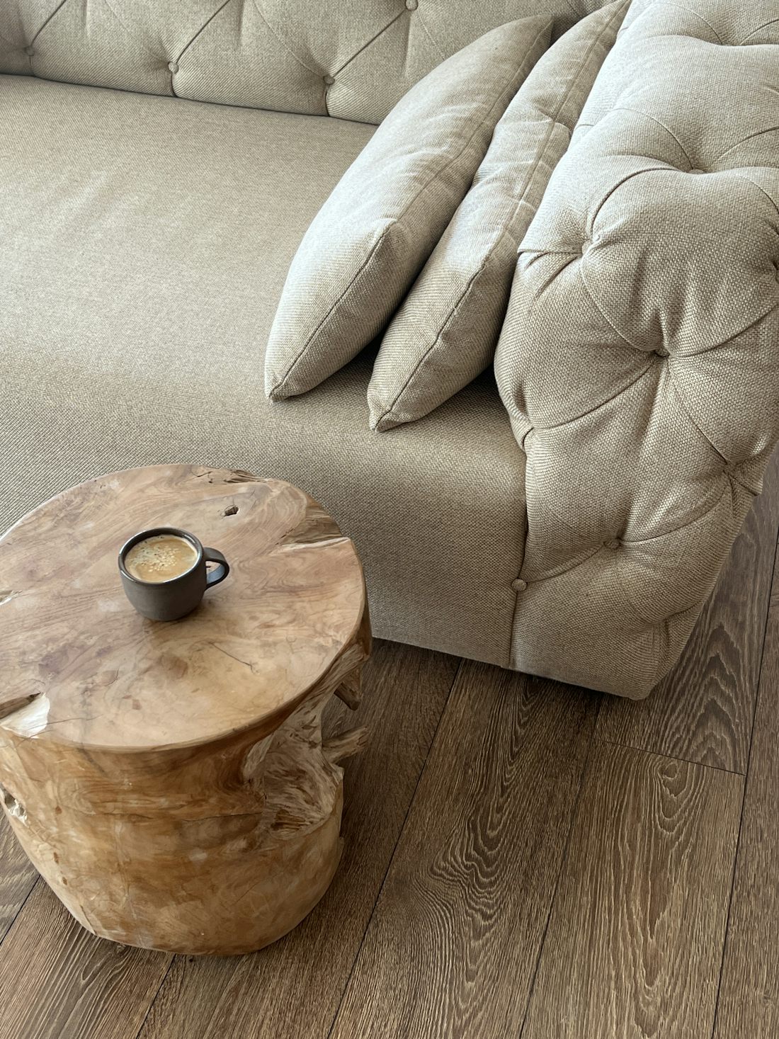 A cup of coffee standing on the bare timber top of a round side table, beside the corner of a buttoned oatmeal linen sofa on a dark oak floor.
