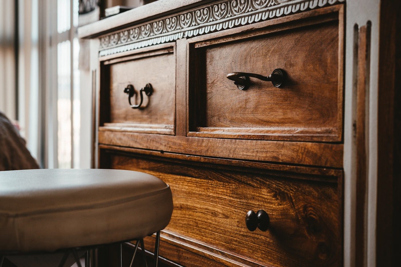 Warm walnut drawer fronts with cast handles, and the pale leather seat of a stool standing in front of them in window light.